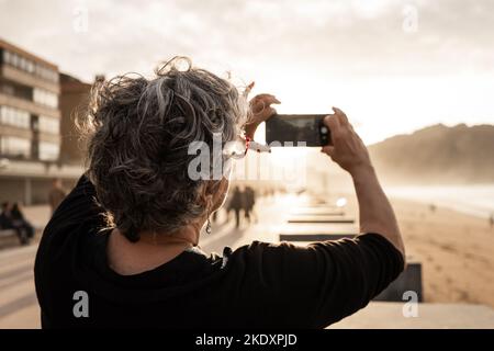 Une femme sénior anonyme qui se rend à la plage et à la mer pour prendre ses cheveux gris tout en passant du temps sur les berges au coucher du soleil sur le complexe Banque D'Images