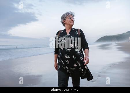 Femme âgée avec des cheveux gris bouclés inhalant de l'air frais tout en passant du temps sur la plage près de la mer en agitant la journée trouble terne Banque D'Images