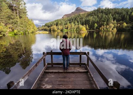 Vue arrière d'une touriste femelle méconnaissable se trouvant sur un quai en bois près d'un lac calme contre la forêt de Glencoe Lochan et le pic de montagne le jour nuageux à SCO Banque D'Images