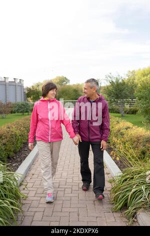 Tout le corps heureux homme et femme d'âge moyen tenant les mains et regardant l'un l'autre tout en se promenant sur le chemin pavé dans le parc Banque D'Images