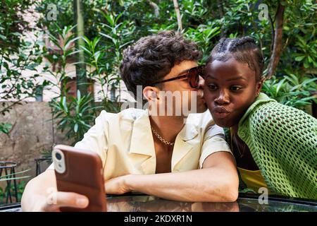 Un jeune homme élégant embrasse une petite amie noire qui se repose sur une table et prend son selfie via un smartphone tout en étant assise à table pendant un séjour romantique Banque D'Images