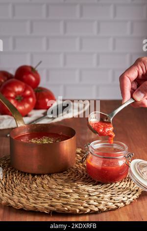 Personne méconnaissable utilisant une cuillère pour remplir le pot en verre de sauce tomate contre le mur de brique blanche dans la cuisine à la maison Banque D'Images
