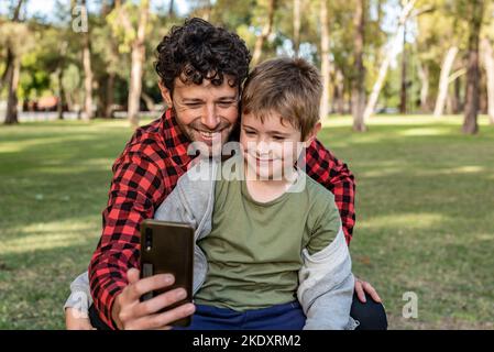 Homme positif prenant autoportrait sur smartphone avec heureux préteen fils tout en étant assis sur l'herbe dans la forêt en été Banque D'Images