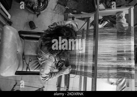 Vue de dessus en noir et blanc d'une travailleuse anonyme avec des cheveux bouclés assis sur une chaise confortable près du métier à tisser et du tapis de tissage dans l'atelier Banque D'Images