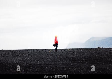 Voyageur dans une veste rouge debout sur une plage de sable noir en Islande Banque D'Images