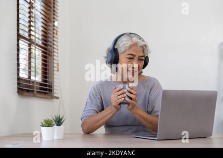 Femme âgée utilisant un ordinateur portable tout en portant un casque à la maison - concept de mode de vie et de technologie joyeux pour les personnes âgées Banque D'Images