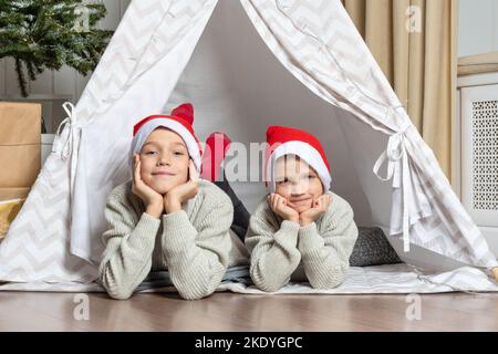 Noël en famille. Deux frères dans un chapeau de Père Noël, allongé dans une tente pour enfants dans la chambre, attendant impatiemment Noël et cadeaux. Les enfants sont waiti Banque D'Images