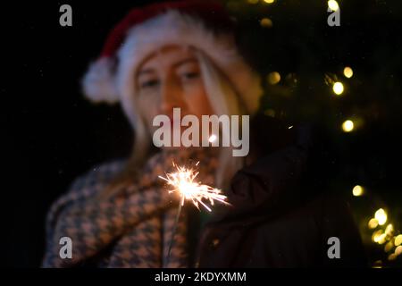 Une jeune femme triste dans un chapeau de Père Noël regarde un sparkler dans ses mains sur le fond des décorations de Noël de la ville la nuit Banque D'Images