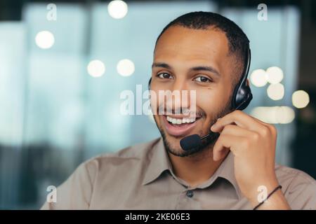 Portrait un professionnel de l'assistance technique souriant et heureux avec un casque pour les appels vidéo, un homme afro-américain regardant la caméra de proximité, travaillant à l'intérieur d'un centre d'appels de bureau moderne, un service d'assistance clientèle en ligne. Banque D'Images