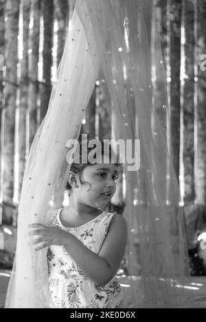 Photo verticale en niveaux de gris d'une petite fille sud-asiatique assise sur le lit et se cachant derrière un voile rose Banque D'Images