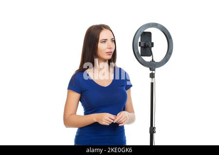 La belle fille blogger communique avec les abonnés de son canal en utilisant un smartphone et un anneau lumineux. Isolé sur un fond blanc. Banque D'Images
