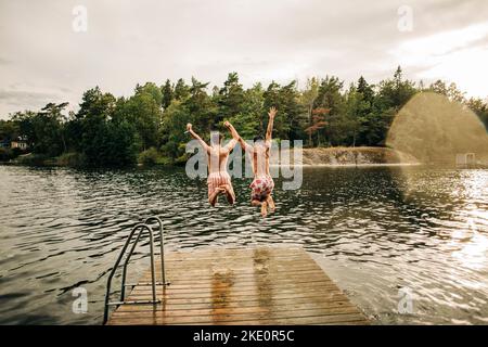 Des amis masculins insouciants avec des bras levés sautant dans le lac pendant les vacances Banque D'Images