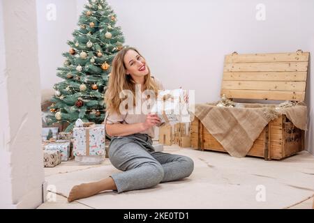 Élégante et moderne, cette femme millénaire est assise à l'arbre de Noël artificiel, tient un cadeau entre ses mains, sourit. Rouge à lèvres sur les lèvres. Ambiance festive et écologique Banque D'Images