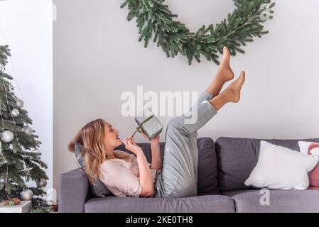 Femme millénaire avec rouge à lèvres en forme de punaise, tenant un cadeau dans les mains, les jambes soulevées sur un canapé gris près de l'arbre de Noël, couronne. Fou Nouveau Banque D'Images