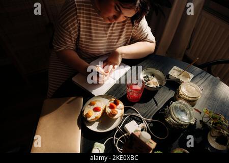 Vue en grand angle de la jeune femme écrivant dans un journal tout en étant assise à table Banque D'Images