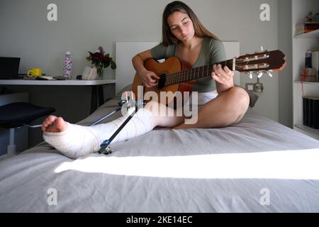 Jeune femme avec jambe cassée couché sur le lit jouant de la guitare Banque D'Images