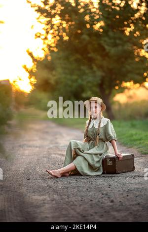 une fille au coucher du soleil est assise sur la route dans un chapeau et une robe vintage Banque D'Images