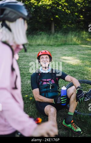 Joyeux cycliste assis sur l'herbe tenant une bouteille d'eau tout en parlant à une amie femelle Banque D'Images