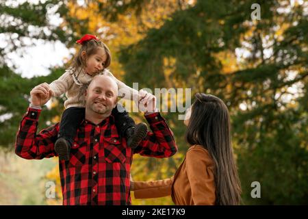 Petite fille mignonne assise sur les épaules de son père dans le parc lors d’un beau jour d’automne et jouant avec sa mère. Ils dépensent fa précieux Banque D'Images