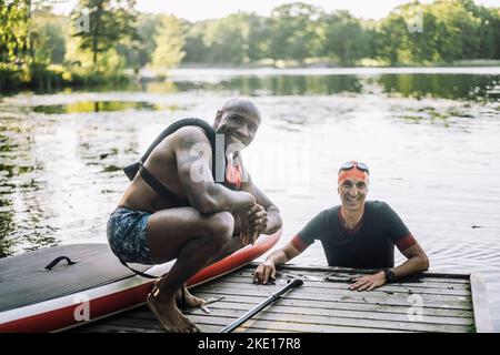 Portrait d'un homme souriant portant un gilet de sauvetage accroupi sur la jetée par un ami mâle dans le lac Banque D'Images