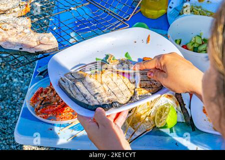 Une femme tenant une fourchette, des tranches d'espadon sur une assiette blanche et un barbecue. Accent sélectif sur une table en plastique, une tranche de fruits au citron et des têtes de crevettes. Banque D'Images