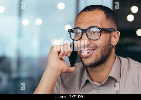 Photo en gros plan d'un homme d'affaires afro-américain souriant et heureux en lunettes parlant gaiement au téléphone avec les clients, un vendeur masculin travaillant dans un immeuble de bureaux moderne. Banque D'Images