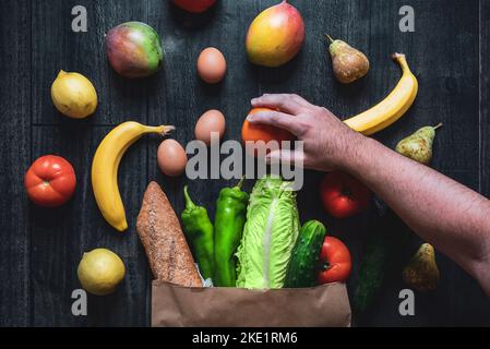 Une main cueillant un fruit d'un sac de shopping rempli de légumes, fruits, oeufs et pain sur un fond de bois. Choix d'aliments sains. Superma en ligne Banque D'Images