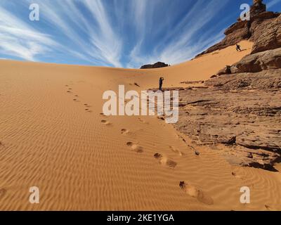 vue à angle bas d'un homme qui filme un autre avec son téléphone, commencez à courir vers le bas d'une dune de sable rocheux avec des empreintes de pas. Ciel bleu ciel nuageux en arrière-plan. Banque D'Images