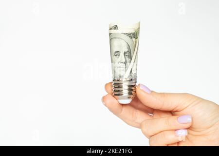 La main d'une femme tient une cartouche d'une ampoule avec des dollars sur fond blanc, le prix de l'électricité, la technologie Banque D'Images