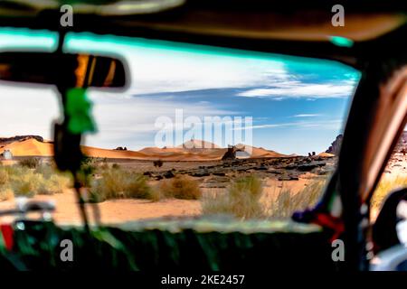 Vue sur le pare-brise depuis l'intérieur d'une voiture des dunes de sable du désert du Sahara, des montagnes rocheuses mesa. Herbes et arbres secs, ciel bleu ciel nuageux coloré. Tadrar Rouge, Dja Banque D'Images