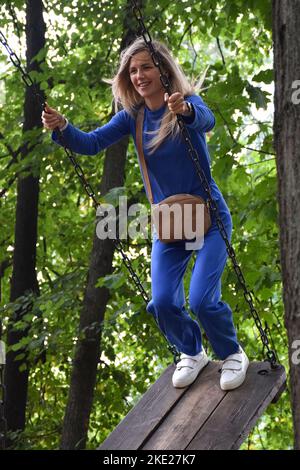 Jolie fille blonde roule sur un carrousel balançoire dans un costume bleu avec un sac à main et sourires Banque D'Images