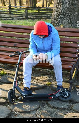 Un adolescent habillé d'un sweat à capuche et d'une veste est assis sur un banc de parc et regarde dans un téléphone portable. À côté de lui se trouve un scooter Banque D'Images