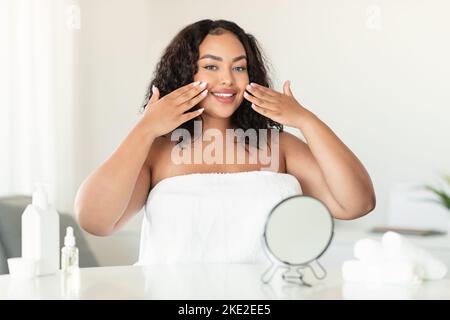 Une jeune femme afro-américaine de plus grande taille touche son visage tout en faisant des soins du visage dans la chambre à coucher, l'espace de copie Banque D'Images