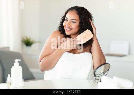 Belle femme positive de corps noir dans une serviette assis dans la chambre, peignant ses cheveux bouclés avec une brosse en bois et souriant Banque D'Images