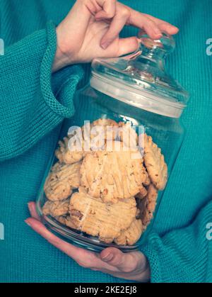 Jeune femme en pull chaud turquois garde le pot en verre avec des biscuits faits maison aux noix dans ses mains, foyer sélectif Banque D'Images