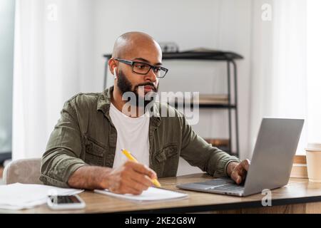Latino-homme indépendant dans les écouteurs à l'aide d'un ordinateur portable et de prendre des notes, ayant la vidéo conférence de la maison, assis au bureau Banque D'Images