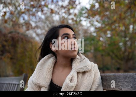 Jeune fille indienne célibataire assise sur un banc dans un parc public qui regarde le ciel. Banque D'Images