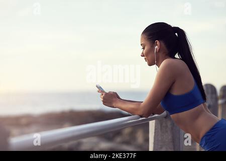 Suivi de ses progrès avec une application de fitness cool. Une jeune femme FIT utilisant son téléphone et des écouteurs pendant un entraînement en plein air. Banque D'Images