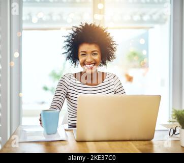 Pas de stress quand vous êtes votre propre patron. Portrait d'une jeune femme joyeuse travaillant sur un ordinateur portable et buvant un café tout en regardant l'appareil photo à la maison. Banque D'Images