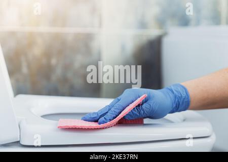 Vue rognée avec gros plan sur les mains de femme gantées de gants de protection en caoutchouc. Femme de ménage nettoyage cuvette de toilette, siège avec tissu rose, essuyer dans la salle de bains ou les toilettes publiques modernes avec carrelage bleu gris Banque D'Images