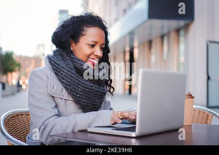 Femme avec un ordinateur portable heureuse de travailler à l'extérieur tout en ayant une réunion en ligne d'affaires, jeune femme souriant et travaillant dans une terrasse de café . Entreprise Banque D'Images