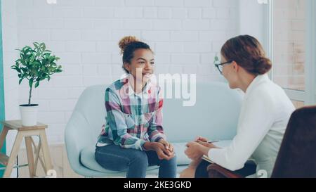 Une jeune fille afro-américaine gaie parle avec un psychologue assis sur un canapé dans un studio moderne de style loft et souriant tandis que le spécialiste l'écoute et prend des notes. Banque D'Images
