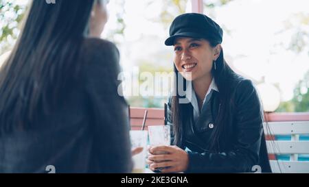 Une fille asiatique heureuse parle à son amie dans un café de rue, puis toaster et trinquer des verres avec des cocktails en appréciant la réunion. Communication, amitié et concept de boissons. Banque D'Images