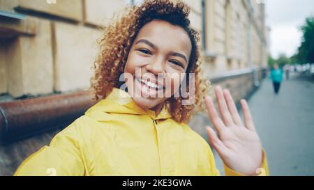 Point de vue prise de jeune femme gaie blogger regardant la caméra, parlant et agitant main enregistrement vidéo pour son vlog en ligne dans l'Internet debout à l'extérieur dans la rue. Banque D'Images