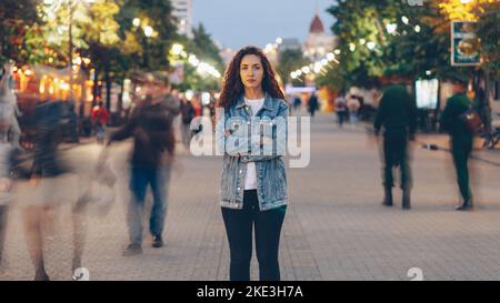 Portrait d'une jeune femme élégante fatiguée de la hâte habituelle se tenant dans la rue parmi les gens qui se précipitent et regardant l'appareil photo. Concept de temps, de jeunesse et de société moderne. Banque D'Images