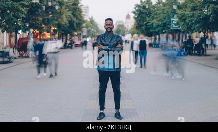 Portrait d'un homme afro-américain heureux portant un Jean élégant et une veste en cuir debout seul dans la rue en centre-ville, souriant et regardant l'appareil photo avec la foule en mouvement. Banque D'Images