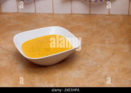 Une élégante assiette carrée profonde remplie d'une crème végétale de carottes et de potiron et de persil sur un comptoir rustique en pierre est dans une cuisine carrelée. Homem Banque D'Images