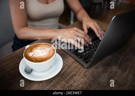 Une femme méconnaissable s'assoit à une table dans un café avec une tasse de café et travaille sur un ordinateur portable Banque D'Images