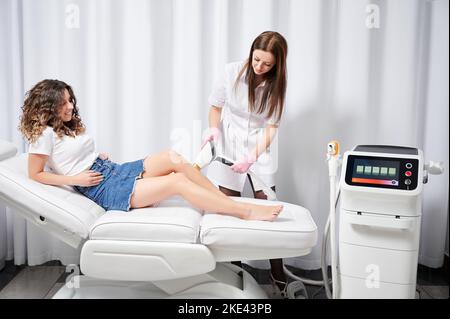 Jeune cosmétologue exécutant la procédure d'enlèvement de laser de cheveux indésirables sur les jambes du client. Méthode efficace d'enlèvement de cheveux de laser pour se débarrasser des cheveux indésirables pour toujours. Banque D'Images