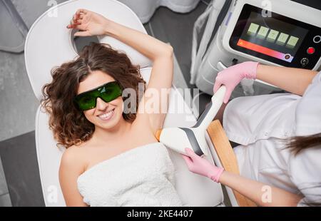 Vue de dessus de la femme joyeuse dans des lunettes de protection couchée sur la chaise de jour et souriant tandis que cosmologiste enlever les cheveux de l'aisselle femelle avec dispositif laser. Esthéticienne exécutant la procédure d'épilation au laser. Banque D'Images